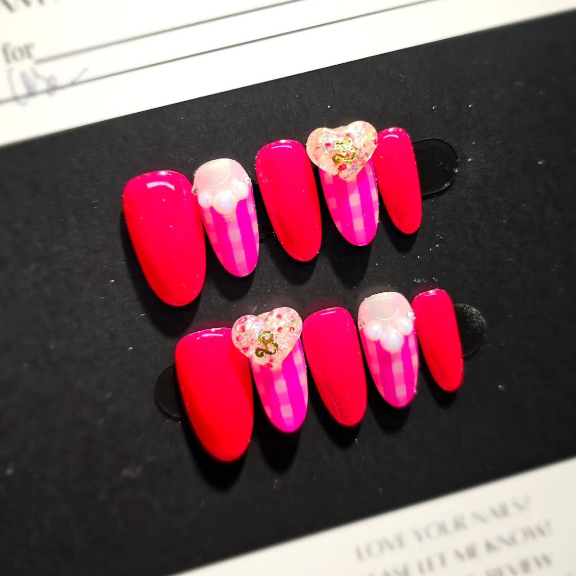 Set of pink and red press-on nails with heart designs on a black surface.